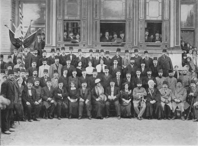 This is a formal photo of a group of
adults in front of a building with windows. There are about four rows of
individuals, those in front seated and those in back standing, plus some
behind in open windows. A substantial majority are men, but there are some women.
Almost all are formally dressed, the men generally in long coats with fez hats.
There are about seventy-five people total