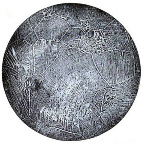 Circular
micrograph showing a Widmanstätten-like microstructure — needle-like
or plate-like crystalline patterns arranged within irregular grain
boundaries, typical of certain metal alloys viewed under a microscope.