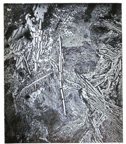 Micrograph
showing a complex, dense metal microstructure with needle-like and
rod-like crystalline formations at various angles, along with wavy
grain patterns.